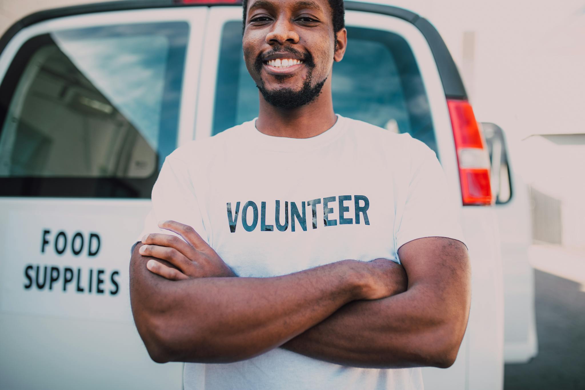 African American volunteer smiling in front of food supplies van with arms crossed, promoting community service.